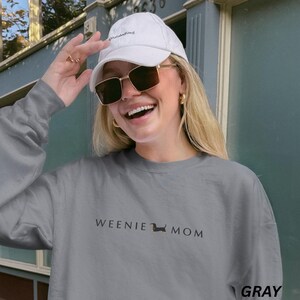 Puede incluir: Sudadera gris con el texto "WEENIE MOM" y una ilustraci&oacute;n de perro salchicha. La sudadera es de color gris liso. Tambi&eacute;n se ven una gorra de b&eacute;isbol blanca y gafas de sol.