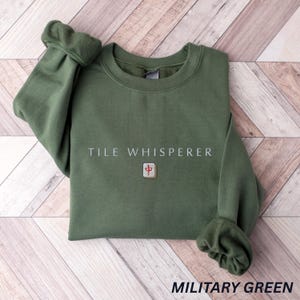 May include: Military green sweatshirt with the text "TILE WHISPERER" and a small graphic. The sweatshirt is a solid color with a crew neck and long sleeves. The sleeves are cuffed at the wrists.
