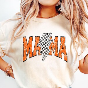 K&ouml;nnte beinhalten: Ein wei&szlig;es T-Shirt mit einer orangefarbenen Grafik, auf der "MAMA" mit einem karierten Blitz in der Mitte steht.