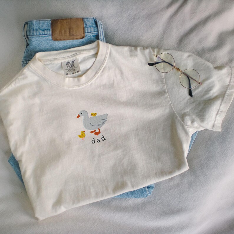 May include: Cream-colored t-shirt with a duck and two ducklings graphic, the word "dad" below. The shirt is paired with light blue jeans and round-framed glasses. The shirt is made of a soft material.