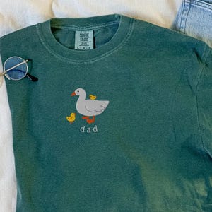 May include: A teal Comfort Colors t-shirt with a cartoon goose and ducklings embroidered on the front. The word "dad" is embroidered below the goose. Round eyeglasses are on the left side of the shirt.
