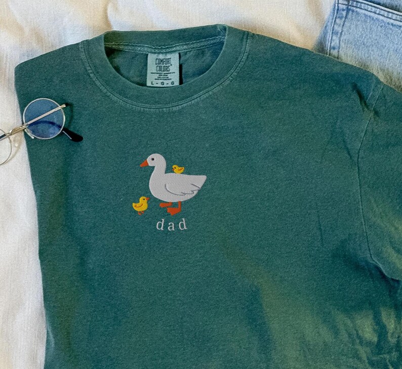 May include: A teal Comfort Colors t-shirt with a cartoon goose and ducklings embroidered on the front. The word "dad" is embroidered below the goose. Round eyeglasses are on the left side of the shirt.