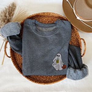 May include: A dark gray sweatshirt with a ghost holding a cup of iced coffee and a small pumpkin. The sweatshirt is laying on a woven basket.