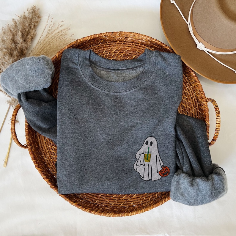 May include: A dark gray sweatshirt with a ghost holding a cup of iced coffee and a small pumpkin. The sweatshirt is laying on a woven basket.