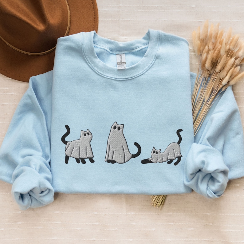 K&ouml;nnte beinhalten: Ein hellblaues Sweatshirt mit einem Design aus drei Katzen, die als Geister verkleidet sind. Die Katzen sind in verschiedenen Posen, eine steht, eine sitzt und eine liegt. Das Design ist in Schwarz und Grau.