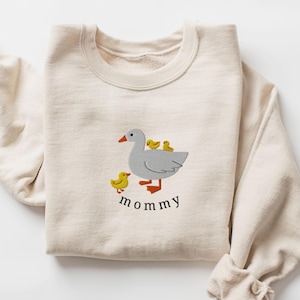 May include: A cream-colored sweatshirt with an embroidered design of a gray goose with three yellow chicks. The text "mommy" is embroidered below the goose.