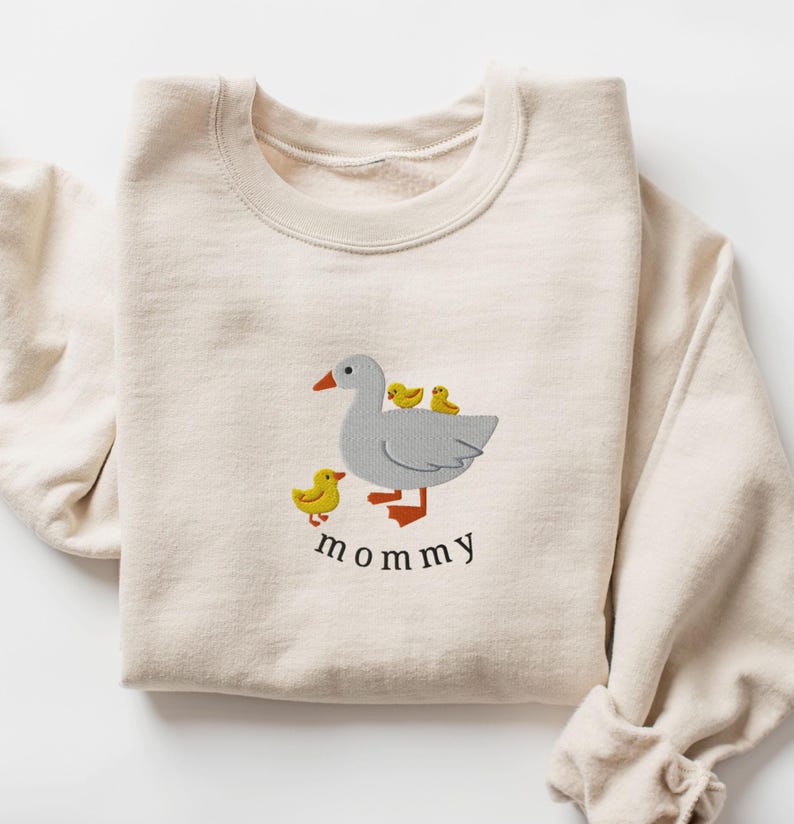 May include: A cream-colored sweatshirt with an embroidered design of a gray goose with three yellow chicks. The text "mommy" is embroidered below the goose.