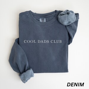 May include: A denim blue sweatshirt with the text "COOL DADS CLUB" in white. The sweatshirt has a crew neck and long sleeves with cuffed wrists. The word "DENIM" is printed in white at the bottom of the image.