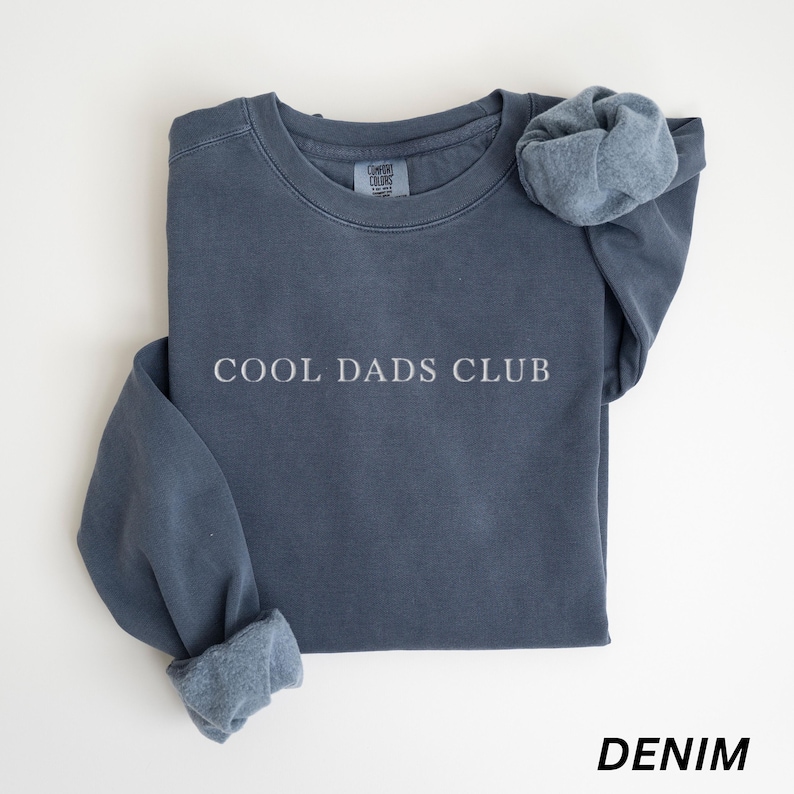 May include: A denim blue sweatshirt with the text "COOL DADS CLUB" in white. The sweatshirt has a crew neck and long sleeves with cuffed wrists. The word "DENIM" is printed in white at the bottom of the image.