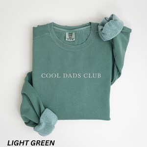 May include: Light green crewneck sweatshirt with the text "COOL DADS CLUB" in white. The sweatshirt is folded with the sleeves draped over the top. The bottom of the image has the text "LIGHT GREEN".