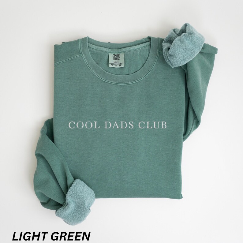 May include: Light green crewneck sweatshirt with the text "COOL DADS CLUB" in white. The sweatshirt is folded with the sleeves draped over the top. The bottom of the image has the text "LIGHT GREEN".