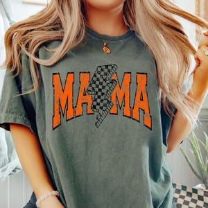 K&ouml;nnte beinhalten: Ein dunkelgr&uuml;nes T-Shirt mit einer abgenutzten orangefarbenen Grafik des Wortes "MAMA" mit einem karierten Blitz in der Mitte.