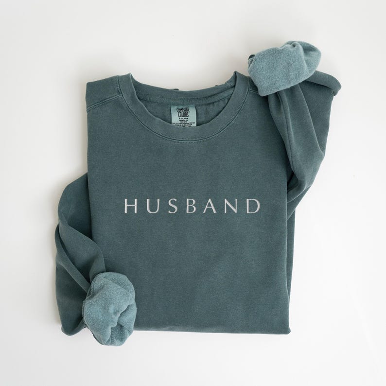 May include: A teal blue sweatshirt with the word "HUSBAND" embroidered in white on the front.