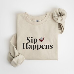 May include: A beige sweatshirt with the text "Sip Happens" and a graphic of a red wine glass with a splash of wine.