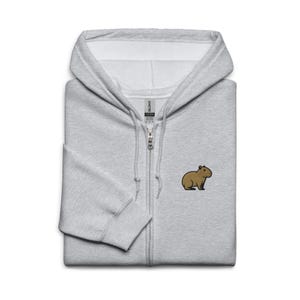 May include: A light gray zip-up hoodie with a small embroidered capybara on the chest. The hoodie has a hood, a full zipper, and ribbed cuffs and hem. The capybara is brown with black accents.
