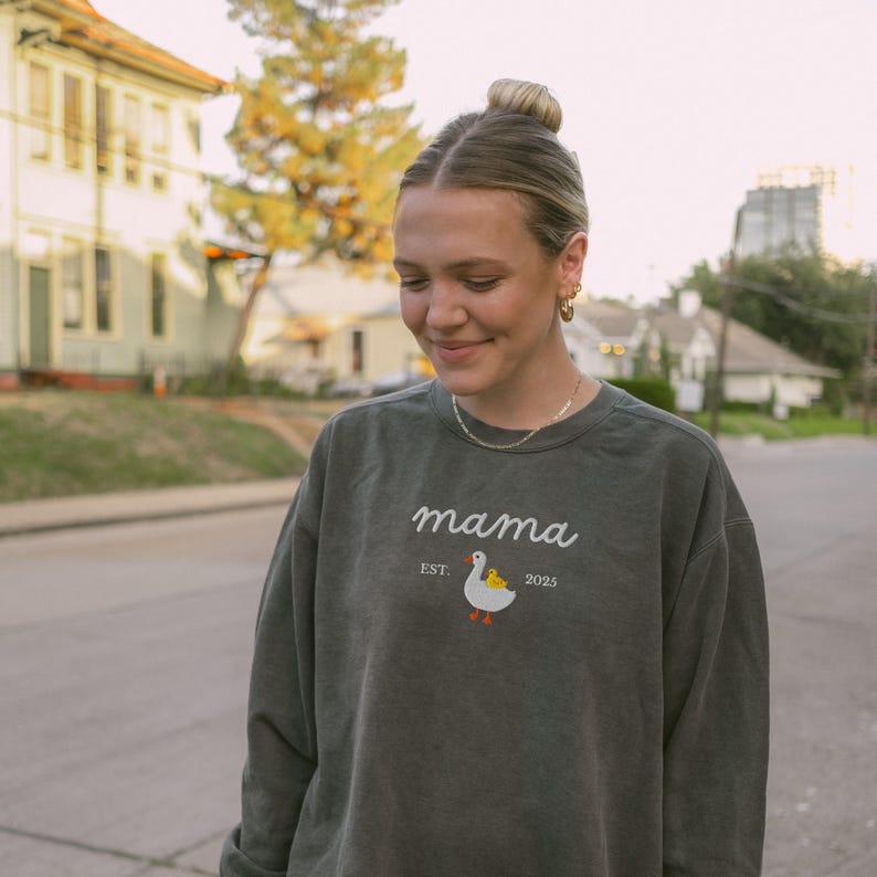 May include: A dark green sweatshirt with the word "mama" in white script. Below the word "mama" is the text "EST. 2025" and a cartoon duck with a yellow chick. The sweatshirt is worn by a person.
