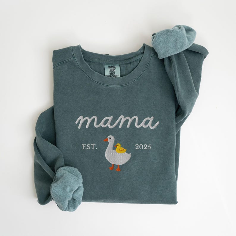 May include: A teal sweatshirt with the word "mama" embroidered in white script. Below the word "mama" is an embroidered goose and duckling design, with the text "EST. 2025". The sleeves are cuffed, and the sweatshirt is made of a soft material.