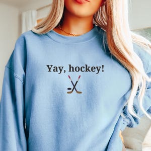 Puede incluir: Una sudadera azul claro con el texto "Yay, hockey!" y un gr&aacute;fico de dos palos de hockey cruzados y un disco.