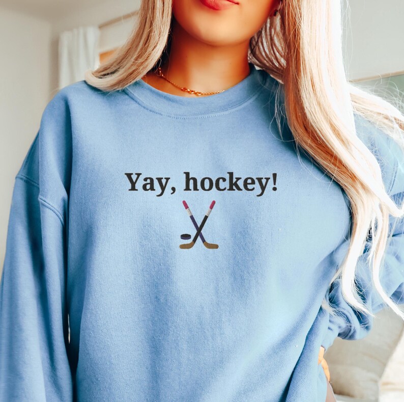 Puede incluir: Una sudadera azul claro con el texto "Yay, hockey!" y un gr&aacute;fico de dos palos de hockey cruzados y un disco.