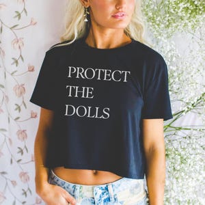 May include: Black crop top with the text "PROTECT THE DOLLS" in white. The shirt is a crew neck style and is paired with blue denim jeans. The background features floral patterns and white flowers.