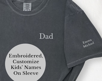 Puede incluir: Una camiseta gris oscuro con la palabra "Papá" bordada en la parte delantera. La manga tiene los nombres "Emma" y "Michael" bordados. El texto "Bordado, Personalizar los nombres de los niños en la manga" está escrito en un círculo en la parte delantera de la camiseta.