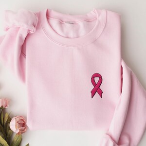 May include: A light pink sweatshirt with a crew neck and long sleeves. A dark pink ribbon is embroidered on the front, near the chest. The sweatshirt is made of a soft material and is likely for casual wear.