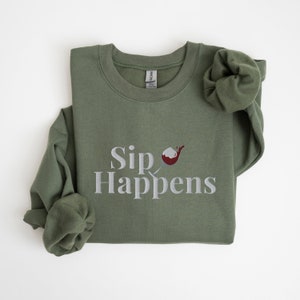 May include: A green sweatshirt with the text "Sip Happens" in white and a red and white cocktail glass graphic.