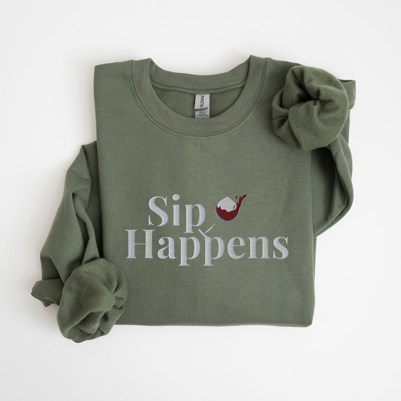 May include: A green sweatshirt with the text "Sip Happens" in white and a red and white cocktail glass graphic.
