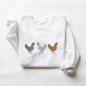 May include: White sweatshirt with a design of three embroidered chickens in a row. The chickens are gray, white, and brown.