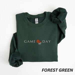May include: Forest green sweatshirt with the text "GAME DAY" and a basketball graphic. The long-sleeved crewneck top is a solid color and has a soft, comfortable appearance. The sweatshirt is a casual style.