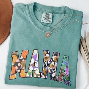 Puede incluir: Una camiseta azul verdosa con la palabra "MAMA" impresa en letras grandes y coloridas. Cada letra est&aacute; llena de un patr&oacute;n diferente con tem&aacute;tica de Halloween, que incluye calabazas, fantasmas, girasoles y telara&ntilde;as.
