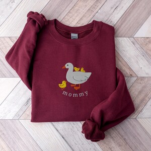 May include: A maroon sweatshirt with an embroidered design of a white duck with three yellow ducklings. The text "mommy" is embroidered below the duck family.