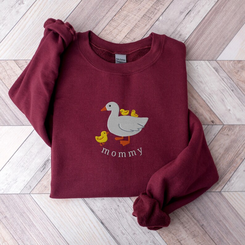 May include: A maroon sweatshirt with an embroidered design of a white duck with three yellow ducklings. The text "mommy" is embroidered below the duck family.