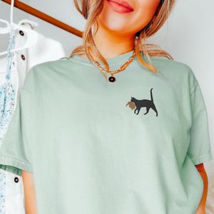 May include: A light green t-shirt with a black cat carrying a brown object on its back. The cat is facing the right side of the image. The t-shirt has a gold chain necklace with a round pendant.