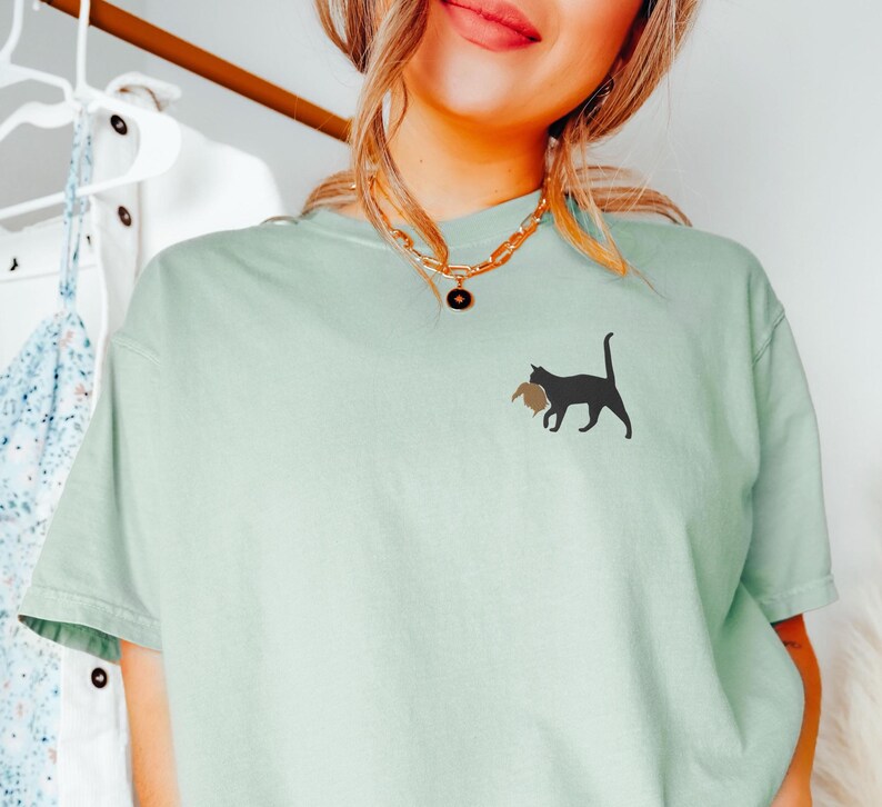 May include: A light green t-shirt with a black cat carrying a brown object on its back. The cat is facing the right side of the image. The t-shirt has a gold chain necklace with a round pendant.