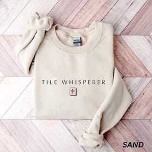 May include: A sand-colored sweatshirt with the text "TILE WHISPERER" and a tile graphic. The sweatshirt has a crew neck and long sleeves with cuffed wrists. The word "SAND" is printed in the bottom right corner.
