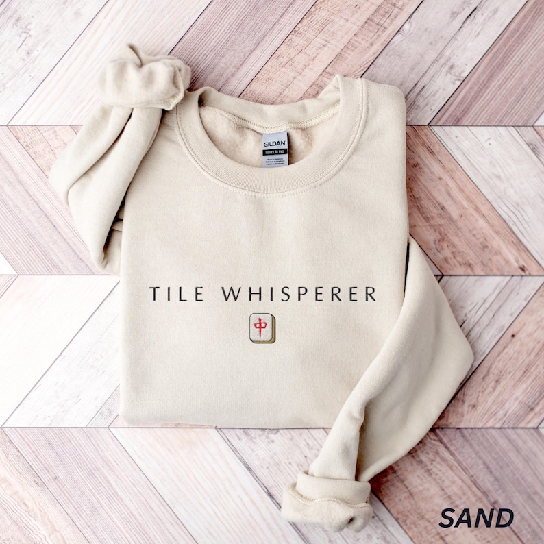 May include: A sand-colored sweatshirt with the text "TILE WHISPERER" and a tile graphic. The sweatshirt has a crew neck and long sleeves with cuffed wrists. The word "SAND" is printed in the bottom right corner.