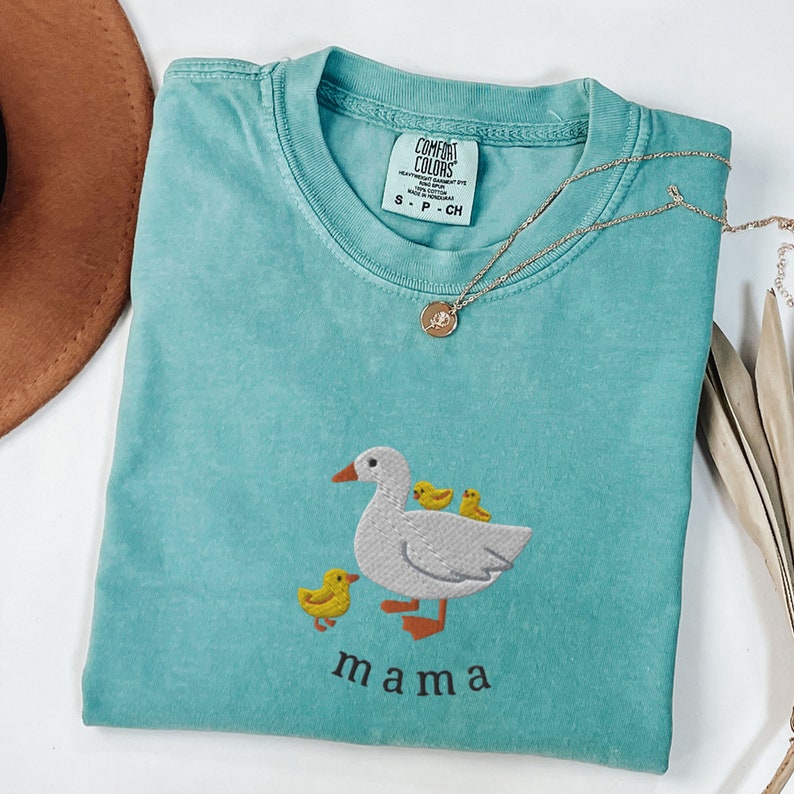 May include: A teal blue t-shirt with an embroidered design of a mama duck and her ducklings. The text "mama" is embroidered below the ducks.