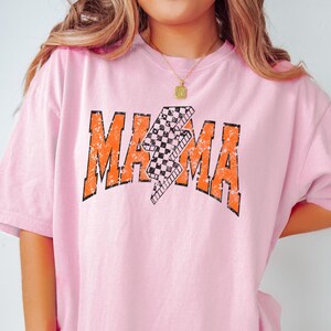 K&ouml;nnte beinhalten: Pinkes T-Shirt mit einem verwaschenen orangenen Grafikdruck des Wortes "MAMA" mit einem karierten Blitzdesign.