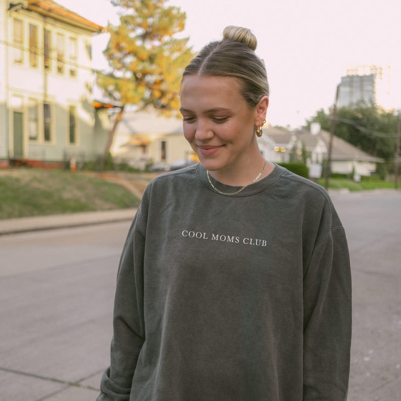 May include: A dark green sweatshirt with the text "COOL MOMS CLUB" in white. The sweatshirt has a relaxed fit and appears to be made of a soft material. The image is taken outdoors.