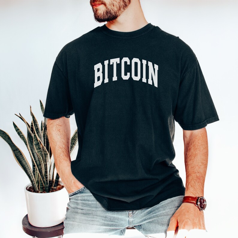 May include: A black t-shirt with the word "BITCOIN" printed in white on the front. The shirt is worn by a person with a beard and is styled with blue jeans.