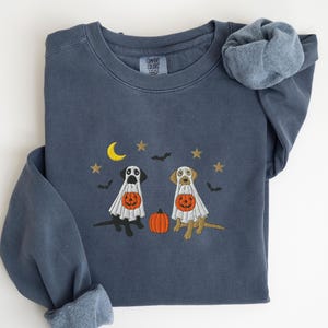 May include: A navy blue sweatshirt with embroidered Halloween-themed dogs. The design features two dogs dressed as ghosts, holding jack-o'-lanterns, with a crescent moon, stars, and bats. The sweatshirt has a Comfort Colors tag.