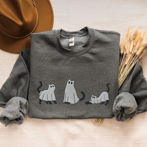 K&ouml;nnte beinhalten: Ein dunkelgrauer Pullover mit einem Design aus drei Geisterkatzen. Die Katzen stehen in einer Reihe, die gr&ouml;&szlig;te in der Mitte und die kleinste rechts. Die Katzen sind alle wei&szlig; mit schwarzen Augen und M&auml;ulern.