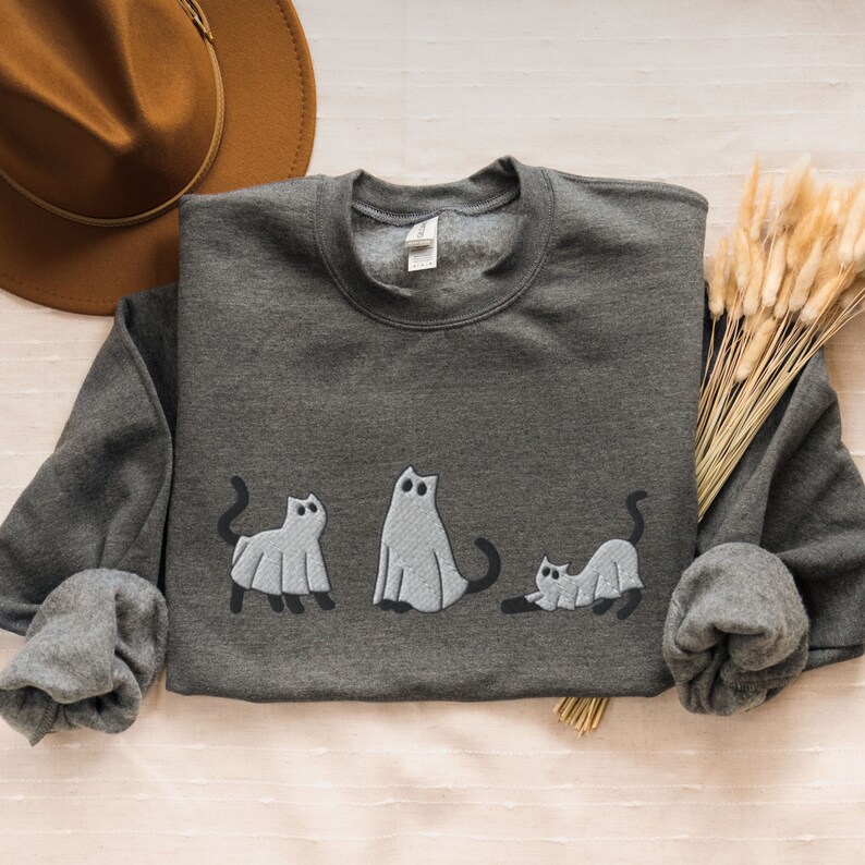 K&ouml;nnte beinhalten: Ein dunkelgrauer Pullover mit einem Design aus drei Geisterkatzen. Die Katzen stehen in einer Reihe, die gr&ouml;&szlig;te in der Mitte und die kleinste rechts. Die Katzen sind alle wei&szlig; mit schwarzen Augen und M&auml;ulern.
