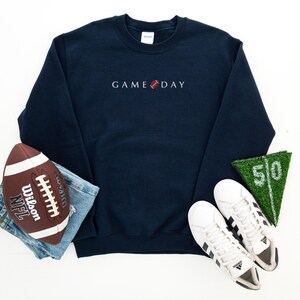 Może przedstawiać: Granatowa bluza z haftem "GAME DAY" w kolorze białym, z grafiką piłki nożnej. Na zdjęciu znajduje się r&oacute;wnież brązowo-biała piłka nożna, niebieskie dżinsy, białe trampki i zielony kawałek murawy z numerem 50.