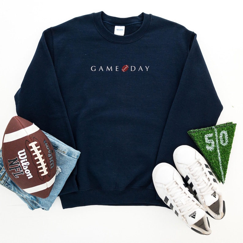 Może przedstawiać: Granatowa bluza z haftem "GAME DAY" w kolorze białym, z grafiką piłki nożnej. Na zdjęciu znajduje się r&oacute;wnież brązowo-biała piłka nożna, niebieskie dżinsy, białe trampki i zielony kawałek murawy z numerem 50.