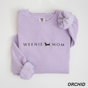 Puede incluir: Sudadera color orqu&iacute;dea con el texto "WEENIE MOM" y una ilustraci&oacute;n de perro salchicha. La camisa de manga larga est&aacute; hecha de un material suave. La palabra "ORCHID" est&aacute; impresa en la parte inferior de la imagen.