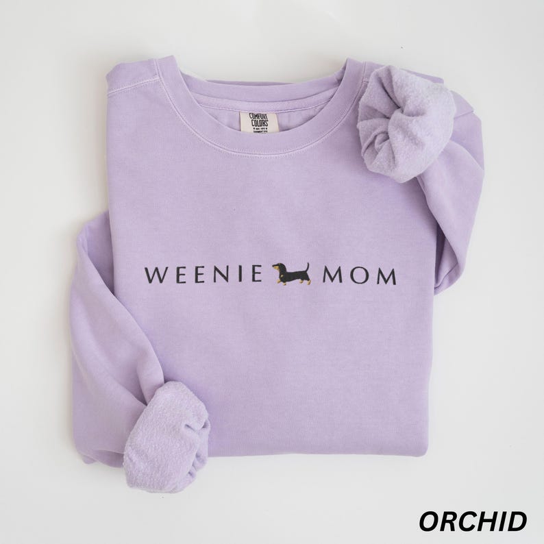 Puede incluir: Sudadera color orqu&iacute;dea con el texto "WEENIE MOM" y una ilustraci&oacute;n de perro salchicha. La camisa de manga larga est&aacute; hecha de un material suave. La palabra "ORCHID" est&aacute; impresa en la parte inferior de la imagen.