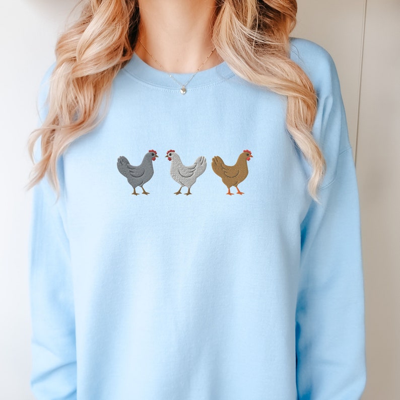 May include: A light blue sweatshirt with an embroidered design of three chickens in a row. The chickens are gray, white, and brown.