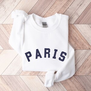 Puede incluir: Sudadera blanca con un gr&aacute;fico "PARIS" azul marino en la parte delantera.